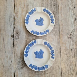 May include: Two white ashtrays with blue decorative elements. Each ashtray has a blue relief image in the center and a blue floral border. The ashtrays are round and have a small indentation for resting cigarettes.