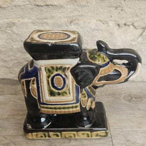 Puede incluir: Figura de elefante de cerámica con cuerpo negro y parte superior cuadrada. El elefante está decorado con patrones azules, verdes y beige. La figura tiene un diseño detallado y está colocada sobre una base rectangular.