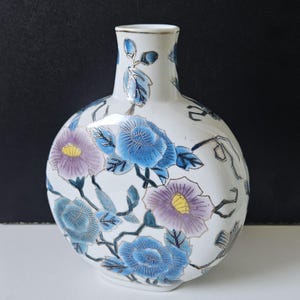 Puede incluir: Jarrón de cerámica blanca con cuerpo redondo y cuello estrecho. El jarrón está decorado con diseños florales azules y morados con detalles dorados. Las flores y hojas están pintadas sobre la superficie del jarrón.