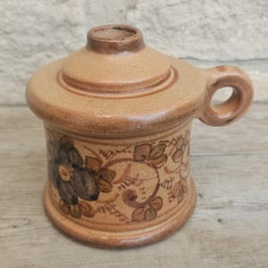 May include: A tan ceramic container with a lid and handle. The container is decorated with a floral design in shades of brown and black. The handle is a small loop. The container is likely for storage or display.