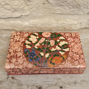 Könnte beinhalten: Rechteckige dekorative Box mit floralem Design. Die Box hat ein zentrales kreisförmiges Design mit einem Baum und Blumen in Rot-, Orange-, Blau- und Weißtönen. Der Hintergrund ist mit rosa Blumen bedeckt.