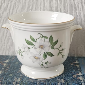 May include: A white ceramic planter with a floral design featuring white flowers and green leaves. The planter has two handles and a gold trim around the rim and base. The planter is approximately 7 inches in diameter.