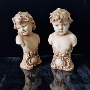 May include: Two ceramic busts of children with curly hair. The busts are in a cream color and have detailed facial features. The bases of the busts are decorated with ornate scrollwork.