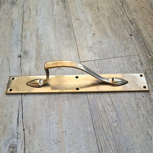May include: A brass door pull with a curved handle and a rectangular backplate. The backplate has decorative pointed ends and several screw holes. The handle is arched and smooth, with a polished finish. The hardware is designed for a door.