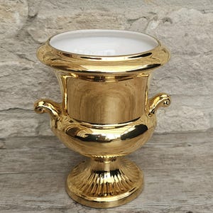 May include: A gold ceramic urn with a white interior. The urn features a wide, rounded rim, two curved handles, and a pedestal base. The glossy surface reflects light. The urn is designed for decorative purposes.