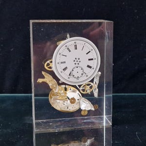 May include: A clear acrylic block containing a vintage watch face and gears. The watch face features Roman numerals and a small second hand dial.