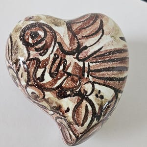 May include: A heart-shaped ceramic decorative object with a hand-painted design. The design features brown, black, and white floral and abstract patterns. The object has a glossy finish and a rounded shape.