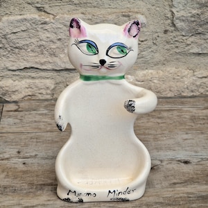 Puede incluir: Un soporte para notas de cerámica color crema con forma de gato, ojos verdes, puntas de las orejas rosas y un collar verde. La figura presenta bigotes y cejas pintados en negro, con el texto "Memo Minder" escrito en negro en la base.