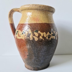 May include: A rustic, handcrafted ceramic pitcher with a handle. The pitcher features a color gradient from light tan at the top to dark brown at the base, with a band of floral-like designs. The pitcher is likely used for serving beverages.