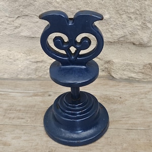 May include: A dark blue metal stand featuring a circular tiered base and a central vertical post supporting a decorative top piece with symmetrical, curved cutouts. The object has a smooth, painted finish with minor signs of wear on the edges.