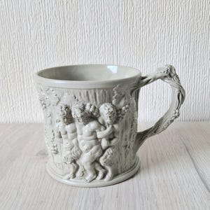 May include: A white ceramic mug with a raised design of three figures in a woodland setting. The mug has a tree trunk handle.