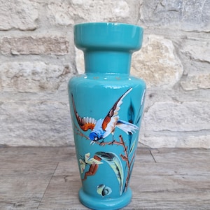 May include: A turquoise ceramic vase with a flared top and base. The vase features a hand-painted design of a bird with blue, brown, and white feathers perched on a branch with leaves. The vase is a decorative home decor item.
