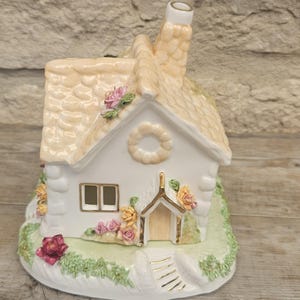 May include: A ceramic cottage figurine featuring a white exterior with gold trim, a textured peach-coloured roof, and a chimney. The base is decorated with sculpted green grass and various colourful floral accents including pink, yellow, and red roses.