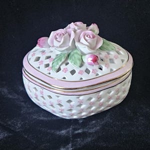 May include: A heart-shaped porcelain trinket box with a white lattice design and pink accents. The lid is adorned with pink rose blossoms and green leaves. The box has a gold trim.