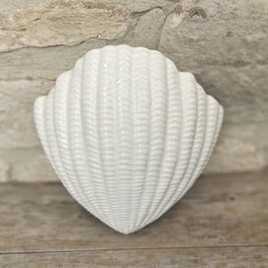May include: A white ceramic seashell-shaped wall decor piece. The shell has a textured surface with detailed ridges and a glossy finish. The decor is set against a neutral background, suggesting a coastal or nautical theme.