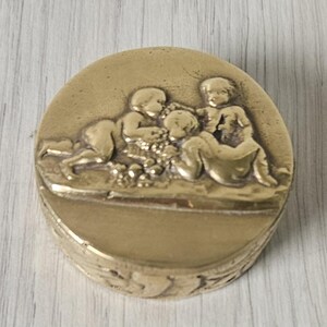 Könnte beinhalten: Eine runde, antike Messingbox mit einem detaillierten Relief von drei Engeln. Die Engel sind in einer spielerischen Szene dargestellt, wobei einer liegt und die anderen sitzen. Die Box hat ein dekoratives Blumenmuster um den Sockel.
