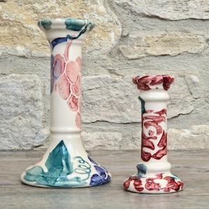 May include: Two ceramic candle holders with a cream-coloured base. The taller holder features a hand-painted grape cluster design in shades of purple, pink, and green. The shorter holder displays a repeating red and blue floral pattern.