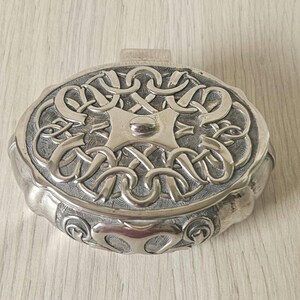 Könnte beinhalten: Eine ovale Schmuckschatulle aus Silber mit einem komplizierten keltischen Knotendesign auf dem Deckel. Die Schachtel hat einen erhabenen, runden Knopf in der Mitte des Deckels.