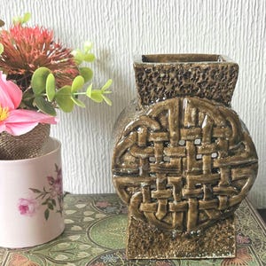 May include: A brown ceramic vase featuring a Celtic knot pattern. The vase has a square top and base, with a rounded body. The vase is approximately 20 cm tall and is displayed with a floral arrangement in a pink and white floral cup.