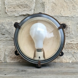 May include: A circular, antique-style light fitting with a clear glass dome and a rusty metal frame. It contains a large, white light bulb. The background is a textured stone wall and a wooden surface.