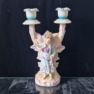 May include: A vintage porcelain double candlestick holder featuring a woman in a traditional dress, holding a fan. The candlestick holders are white with a blue rim. The figurine is light pink, blue, and purple.