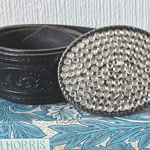 May include: Black leather belt with an oval buckle covered in small, sparkling rhinestones. The belt is rolled up, showing an embossed pattern. The buckle is silver-toned with a twisted rope border.
