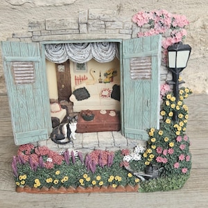 May include: A detailed miniature diorama depicting a charming cottage scene. The scene includes a cat, a sofa, and a table, framed by a stone wall and green shutters. The exterior is decorated with flowers and a lamppost, creating a picturesque, idyllic setting.