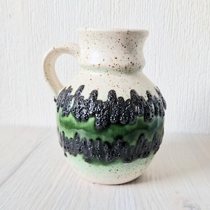 May include: A white ceramic pitcher with a green and black glaze. The pitcher has a rounded body and a handle. The glaze has a textured, speckled look.