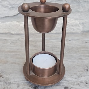 May include: A bronze-colored metal oil burner featuring a circular top with three decorative spheres, a central bowl for oil, and a lower circular base designed to hold a small white tea light candle, supported by three vertical metal rods.