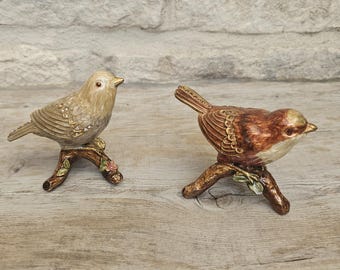 May include: Two bird figurines perched on branches. The left bird is light beige with clear crystal accents on its wings. The right bird is brown with a cream-colored chest and gold-toned wing details. Both birds feature textured feathers and sit on brown branches with small green leaves and pink flowers.