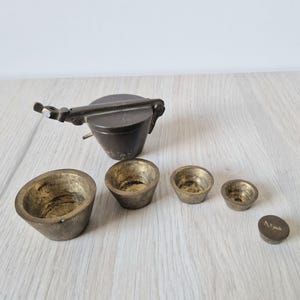 May include: An antique brass scale with a set of small, bowl-shaped weights and a larger, cup-shaped component with a handle. The weights vary in size, and the scale components have a worn, vintage appearance. The items are arranged on a light-coloured wooden surface.