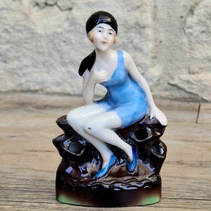 May include: A vintage porcelain figurine of a woman in a blue swimsuit and matching shoes, seated on a dark brown base. The woman has black hair and is posed with one hand on her chest. The base has a green trim.