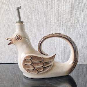 May include: A ceramic bird-shaped oil or vinegar bottle with a spout and handle. The bottle is primarily beige with brown accents on the wings and handle. The bird has an open beak and a metal spout.