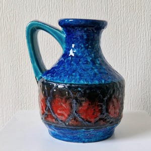 May include: A blue and red ceramic vase with a textured surface and a handle. The vase has a dark blue glaze with a red and black band around the middle.