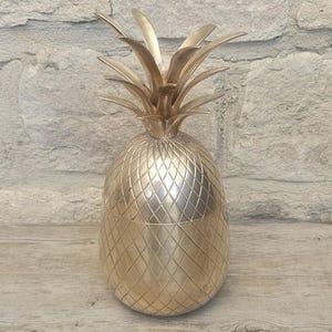 May include: A gold-coloured pineapple-shaped ice bucket. The bucket has a textured, diamond-patterned body and a leafy top. The ice bucket is made of metal and is designed to hold ice and chill beverages.