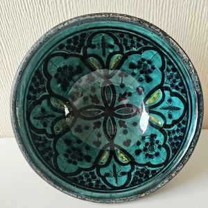 May include: A teal ceramic bowl with a black and yellow floral pattern. The bowl has a dark rim and a glossy finish. The design features a central four-petal flower and repeating floral motifs around the bowl's edge. The bowl is round and has a shallow depth.