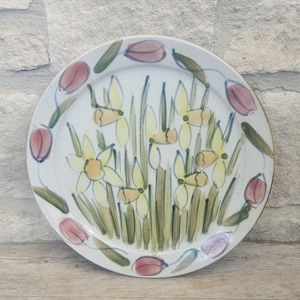 May include: A round ceramic plate with a speckled off-white base. The centre features hand-painted yellow daffodils with green stems and leaves. The rim is decorated with a repeating pattern of pink tulip buds and green leaves painted in a loose style.