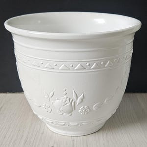 May include: A white ceramic planter with a wide rim and a slightly tapered body. The planter features embossed floral and geometric patterns around the middle. The design includes roses, leaves, and small triangles. The planter is suitable for indoor or outdoor use.