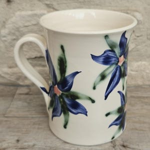 May include: A cream-colored ceramic mug featuring a hand-painted floral pattern with deep blue petals, green leaves, and soft pink centers. The mug has a smooth, cylindrical body with a curved handle on the side, showcasing a glossy finish.