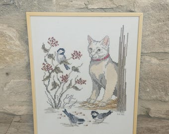 May include: A framed cross-stitch embroidery featuring a white cat with a red collar sitting near a tree trunk. To the left, a bush with red berries and a blue bird is depicted, while two small birds forage on the ground below. The text EH 96 is visible.