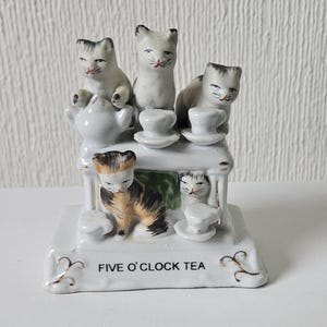 May include: A white porcelain figurine of five cats having tea. The cats are sitting around a table with teacups and a teapot. The base of the figurine is inscribed with the words "FIVE O'CLOCK TEA".