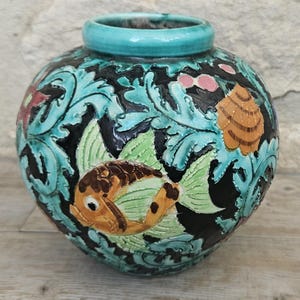 May include: A round ceramic vase with a turquoise rim and a detailed relief design. The vase features a fish, leaves, and floral patterns in shades of orange, green, black, and turquoise. The vase has a bulbous shape.