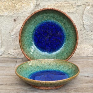 May include: Two handmade ceramic bowls. The bowls feature a blue centre, encircled by a green and brown rim. The larger bowl is behind, and the smaller bowl is in front. The bowls are on a wooden surface.