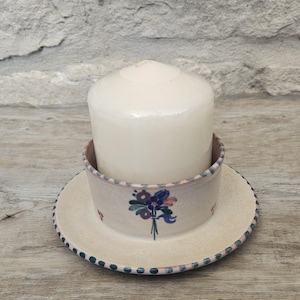 May include: A cylindrical off-white pillar candle sits inside a shallow, circular ceramic dish. The dish features a hand-painted floral motif in blue, purple, and green, with a decorative dotted border along the rim in muted blue and pink tones.