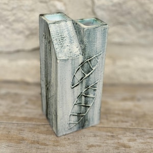 May include: A handcrafted, rectangular vase with a unique design. The vase is painted in shades of gray and white, with leaf-shaped patterns carved into the surface. The top features two square openings, and the overall aesthetic is rustic and artistic.