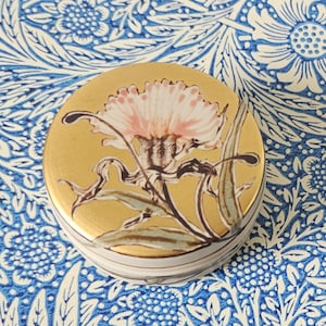 May include: A round trinket box with a gold lid. The lid features a pink and white flower design. The box has a gold rim and a white base with brown stripes. The background is a blue and white floral pattern.