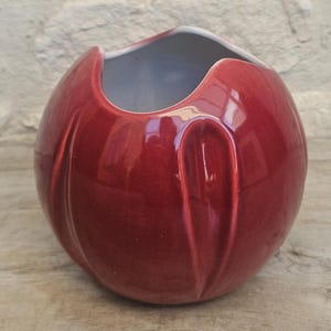 Könnte beinhalten: Eine kugelförmige Keramikvase mit glänzend tiefroter Oberfläche. Die Außenseite weist vertikale Vertiefungen auf, die eine blütenblattartige Form um die Öffnung bilden. Das Innere der Vase ist weiß und der Rand hat ein unregelmäßiges, welliges Design.