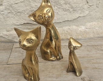 May include: Three brass cat figurines of varying sizes. The cats are posed in different positions, with one sitting, one standing, and one with its head tilted. The figurines have a polished finish and are displayed on a wooden surface.