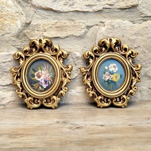 May include: Two oval wall plaques featuring ornate gold-coloured frames with intricate scrollwork detailing. Each plaque contains a central painting of a floral bouquet, one displaying purple and white flowers and the other showing white and yellow roses.