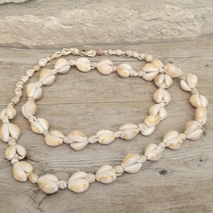 Peut inclure: Un long collier composé de nombreuses petites coquilles de cauris couleur crème enfilées en une boucle continue. Les coquilles présentent des marques naturelles brun clair et beige, avec une finition lisse et polie, disposées en motif régulier.